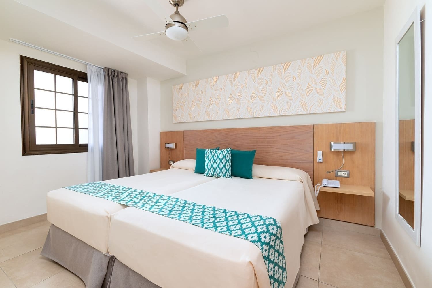 A hotel room with a large bed, a ceiling fan, a window with curtains, a bedside table with a lamp, and a wall-mounted artwork.