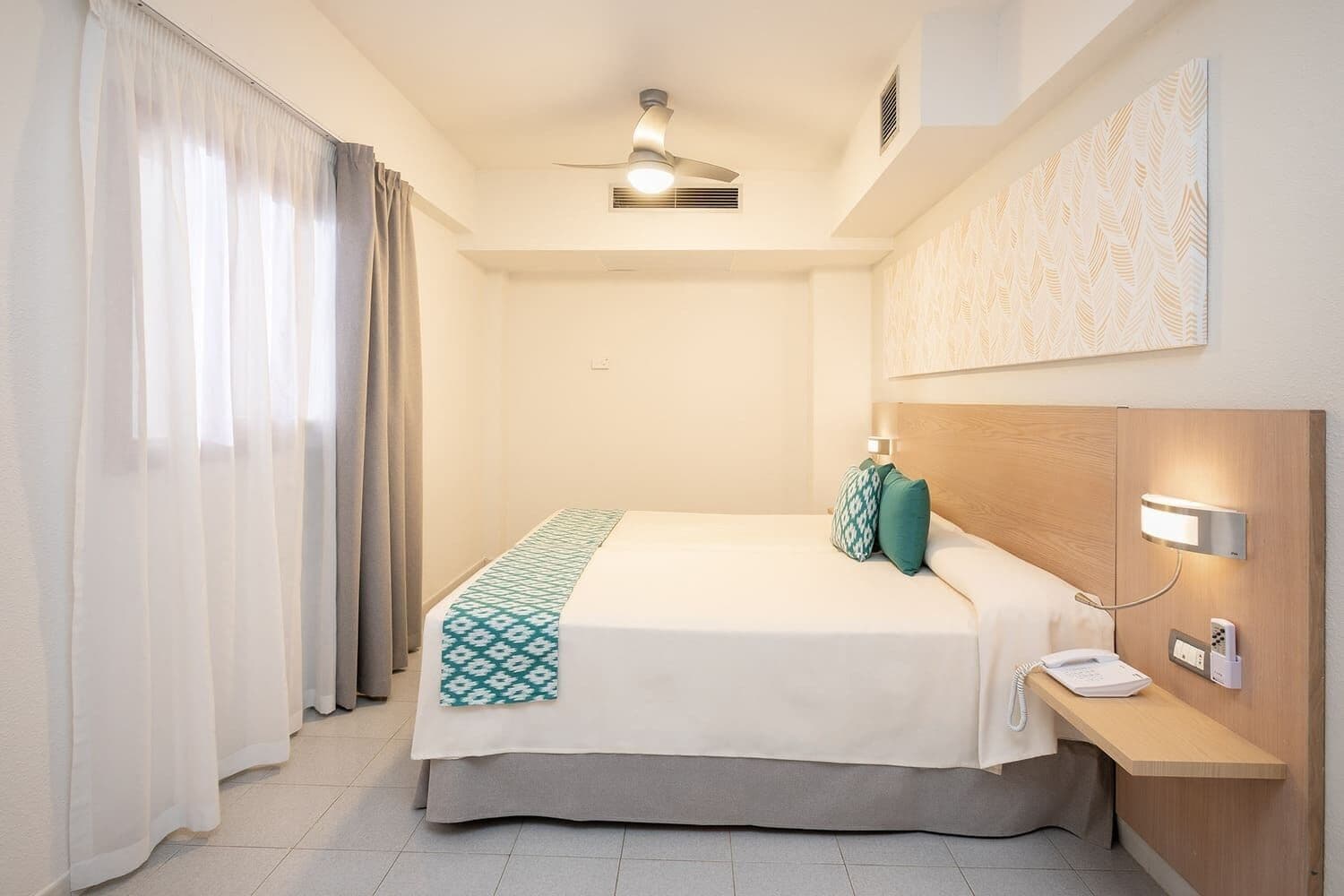 A hotel room with a bed, a bedside table with a phone, a ceiling fan, and a wall-mounted light.