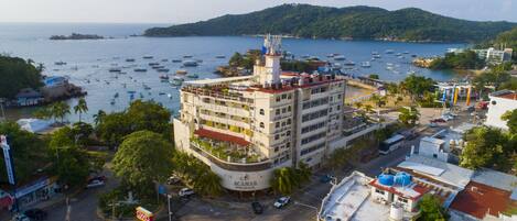 Um hotel com vários andares, piscina no terraço e vista para o mar com barcos.