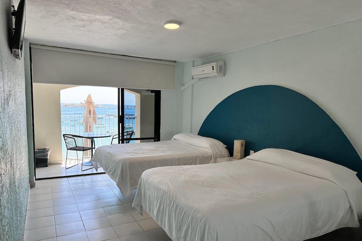 A hotel room with two beds, a balcony with a view of the sea, and a small table and chair.