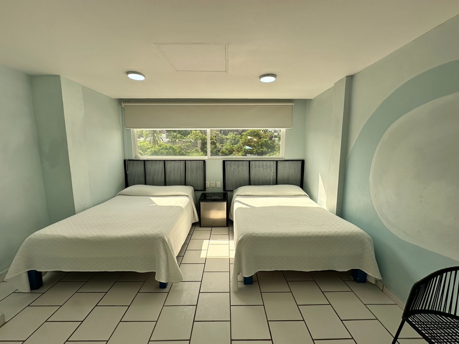 Une chambre d’hôtel avec deux lits simples, une fenêtre avec des rideaux, un radiateur et un sol carrelé.