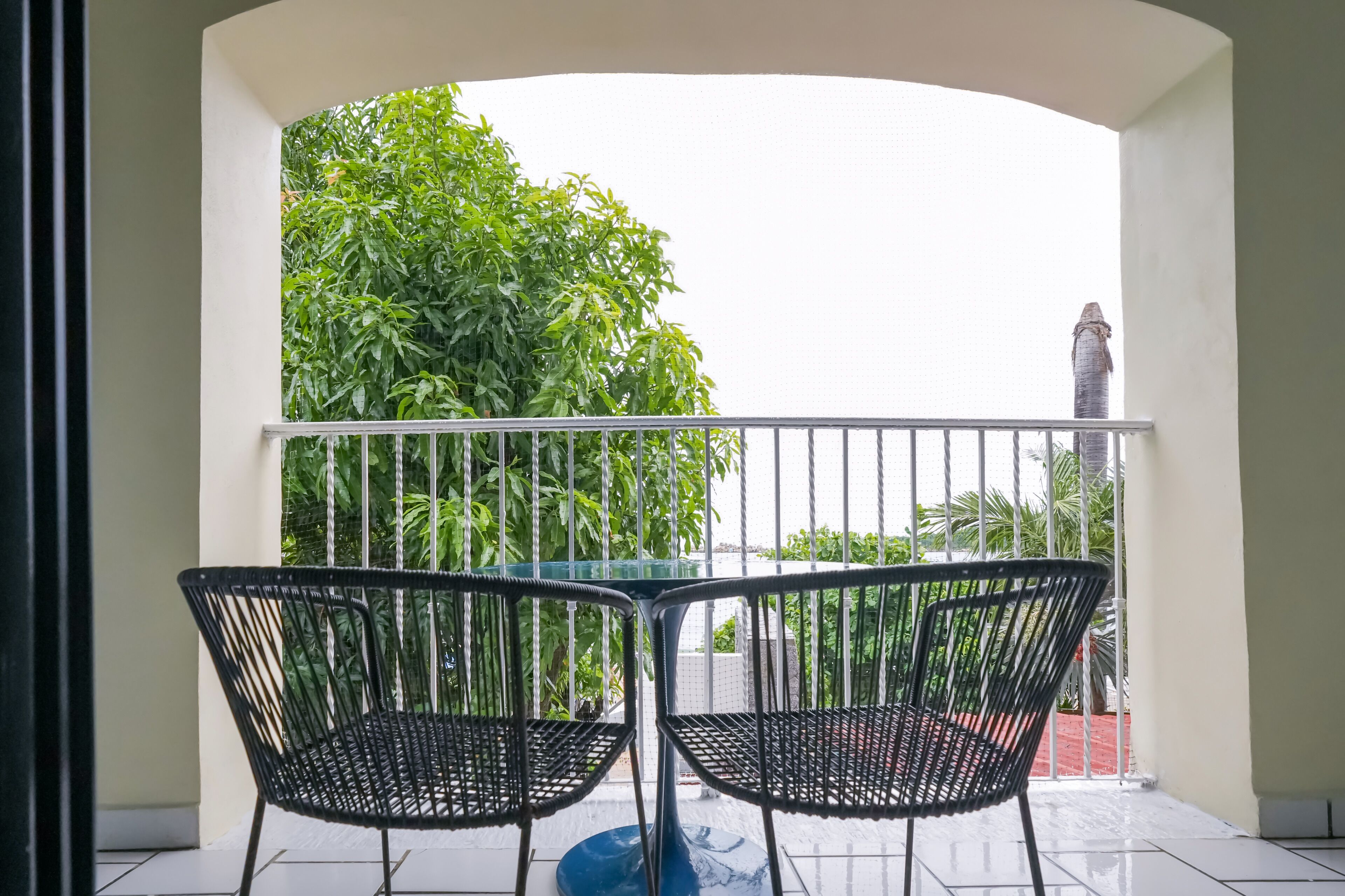 Un balcón con dos sillas y una mesa, rodeado por una barandilla blanca, con vista a follaje verde y una estatua al fondo.