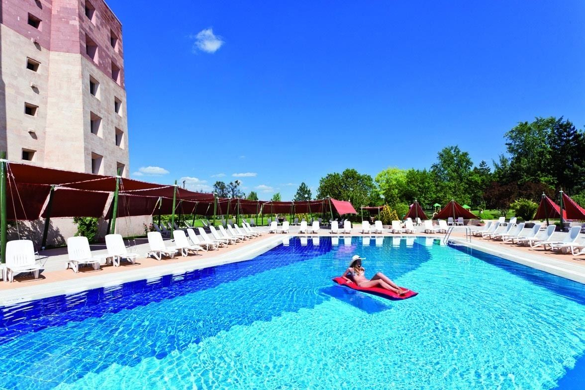 A swimming pool with lounge chairs and umbrellas, surrounded by greenery and a building.