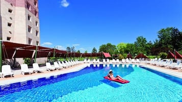 A swimming pool with lounge chairs and umbrellas, surrounded by greenery and a building.