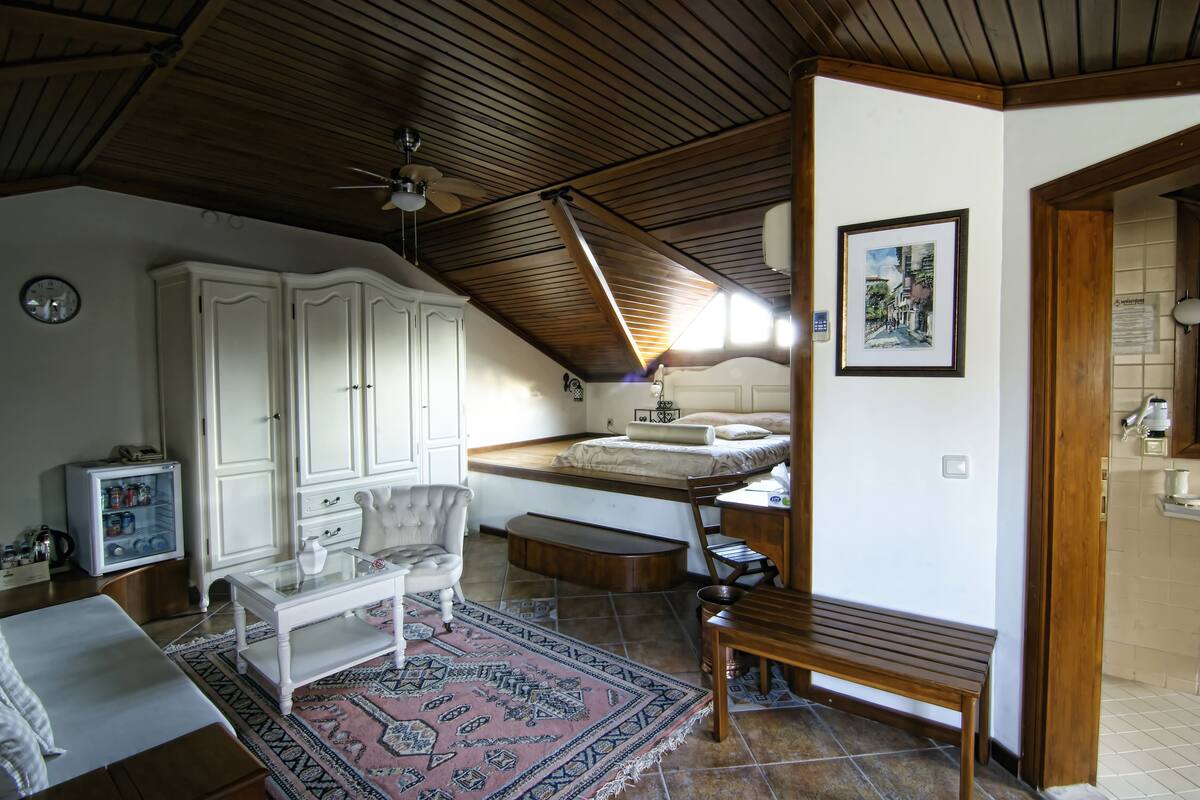 A cozy room with a wooden ceiling, a bed, a white wardrobe, a small table, and a bench.