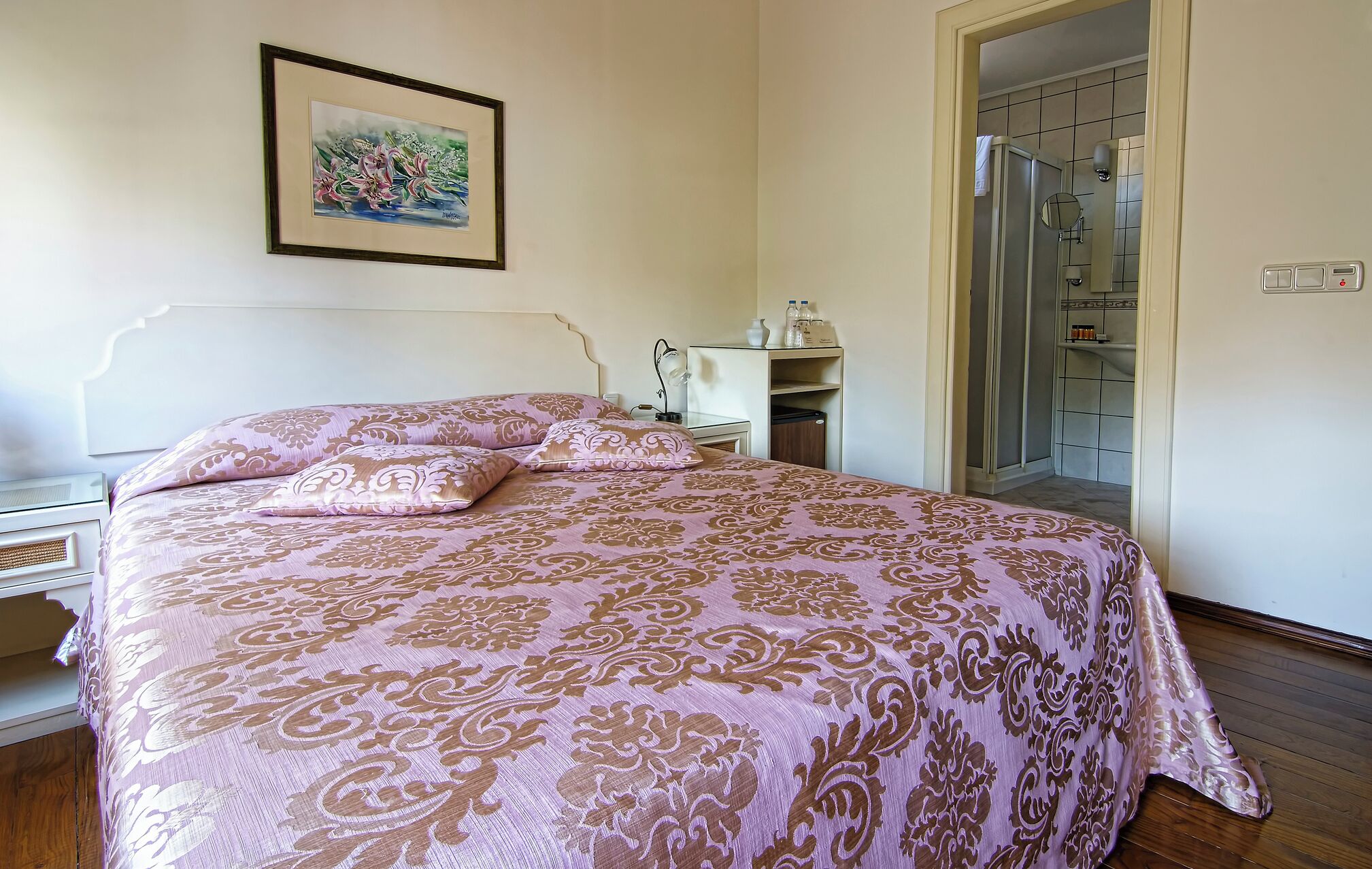 Ein Schlafzimmer mit Bett, Nachttisch, Lampe, einem gerahmten Bild an der Wand und einem durchgegangenen Tür erkennbaren Badezimmer.