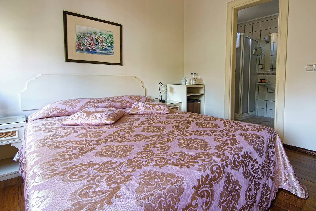 A bedroom with a bed, a bedside table, a lamp, a framed picture on the wall, and a bathroom visible through an open door.