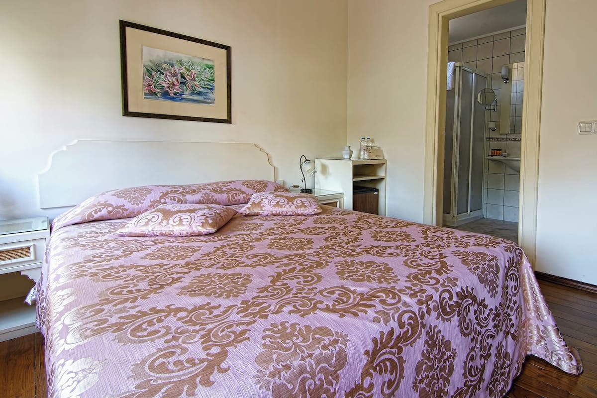 Un dormitorio con cama, mesita de noche, lámpara, un cuadro en la pared y un baño visible a través de una puerta abierta.