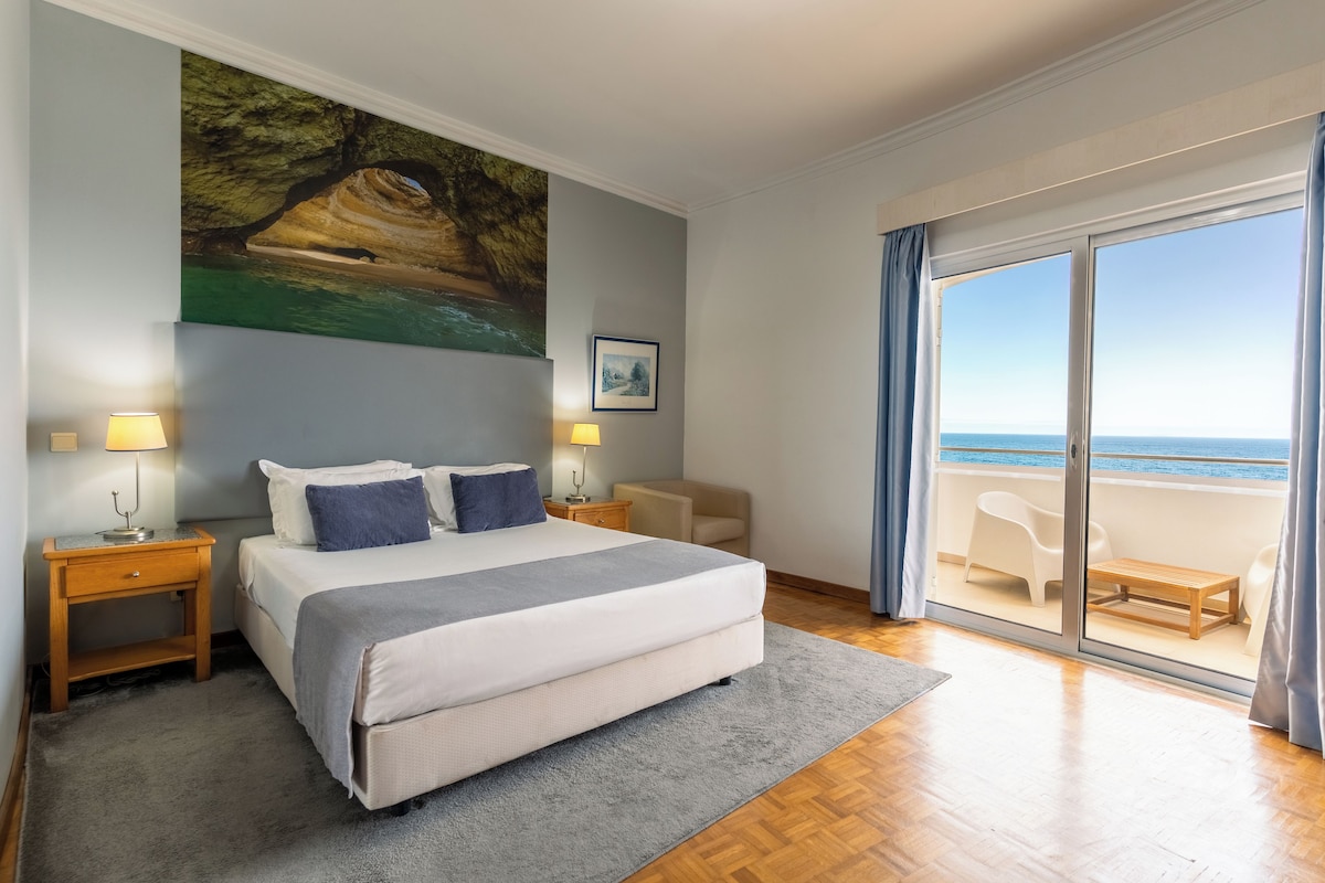 A bedroom with a large bed, a painting of a cave, a window with a view of the sea, and a balcony with outdoor furniture.