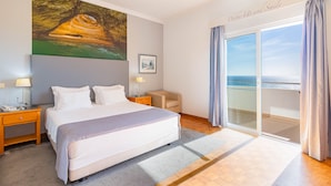 A hotel room with a large bed, a painting of a cave, a desk with a lamp, a chair, and a view of the sea through a sliding door. - Hotel Maritur - Adults Only by Pierre & Vacances (Albufeira)