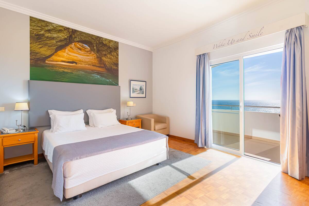 A hotel room with a large bed, a painting of a cave, a desk with a lamp, a chair, and a view of the sea through a sliding door.