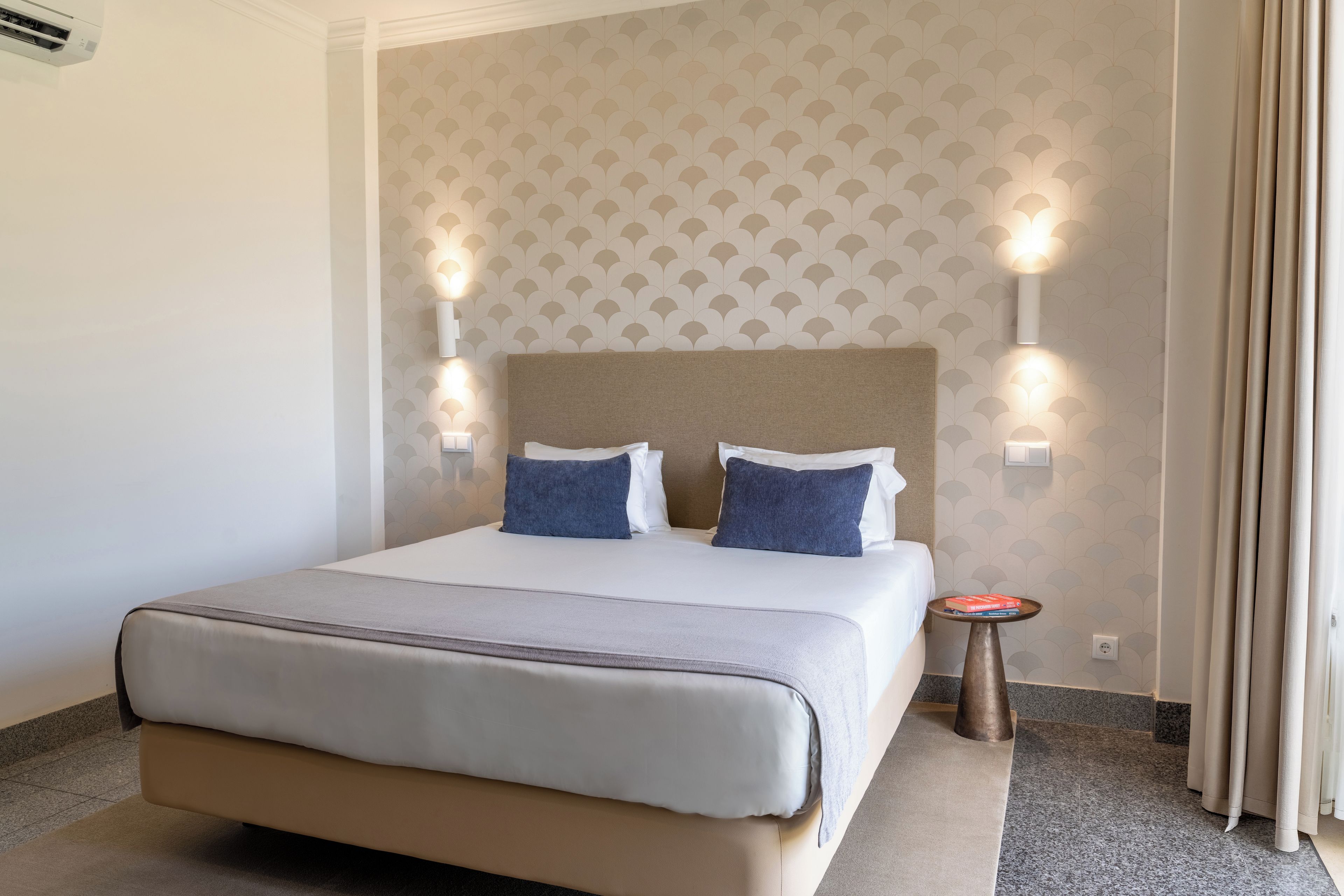 A hotel room with a bed, two wall-mounted lights, a small table, and a curtain.