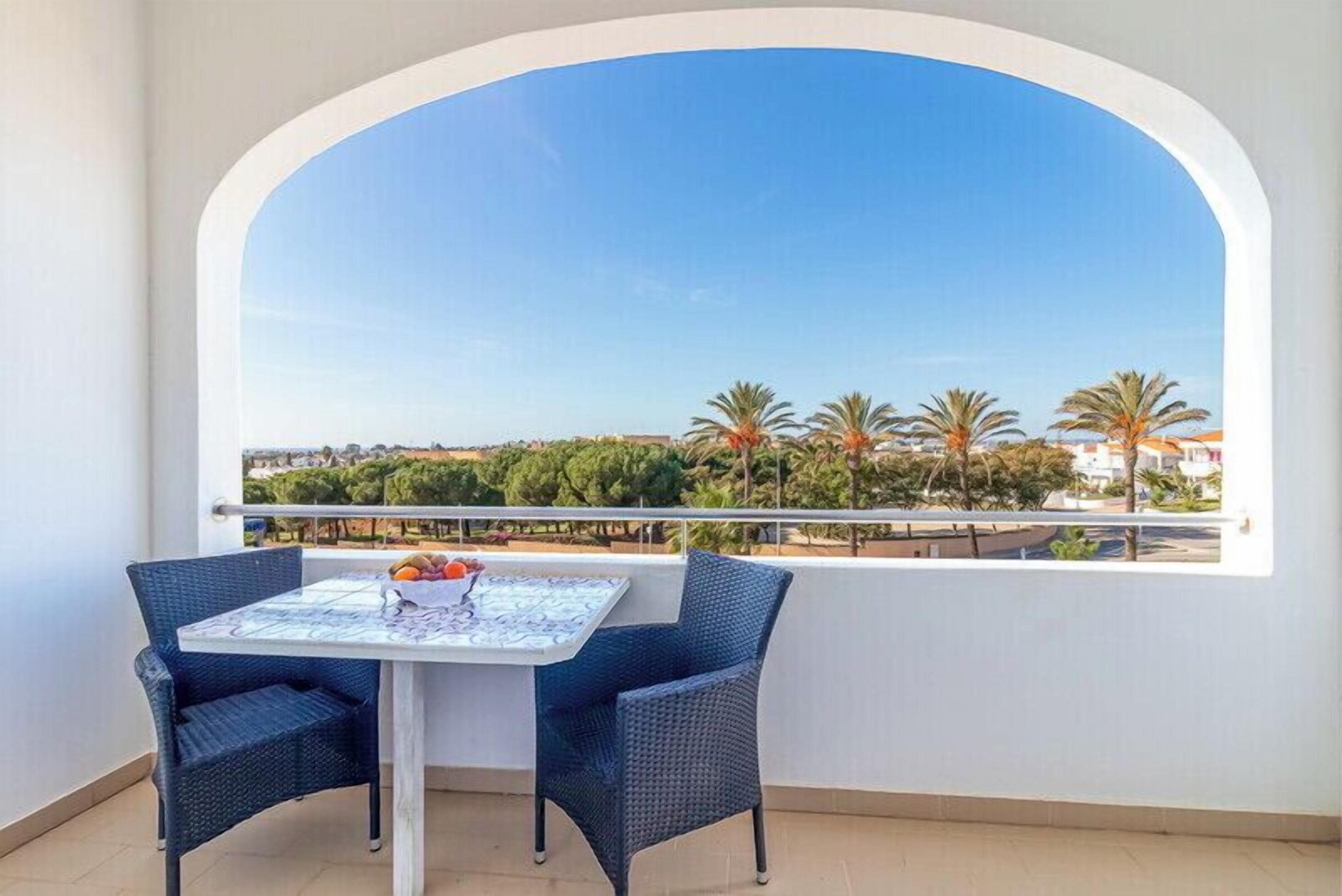 A balcony with a table and chairs, a view of palm trees and buildings, and a clear blue sky.