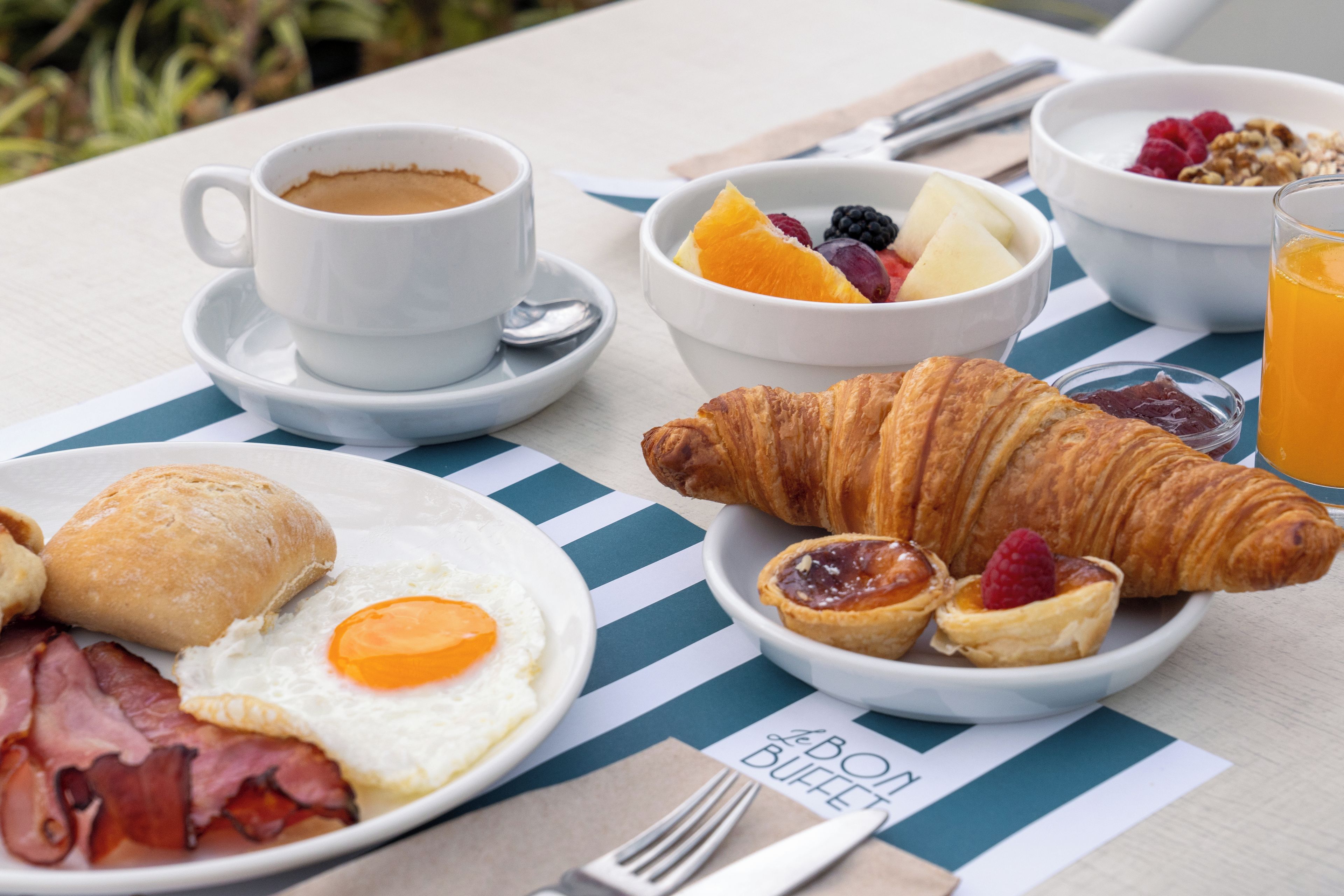 A breakfast table with a cup of coffee, a bowl of fruit, croissants, a plate with eggs and bacon, and a glass of orange juice.