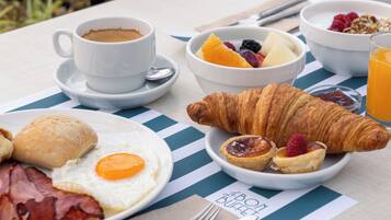 A breakfast table with a cup of coffee, a bowl of fruit, croissants, a plate with eggs and bacon, and a glass of orange juice.