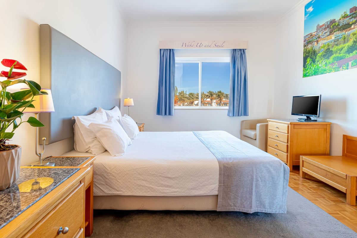 A hotel room with a bed, a desk, a TV, a window with curtains, a plant, and a painting on the wall.