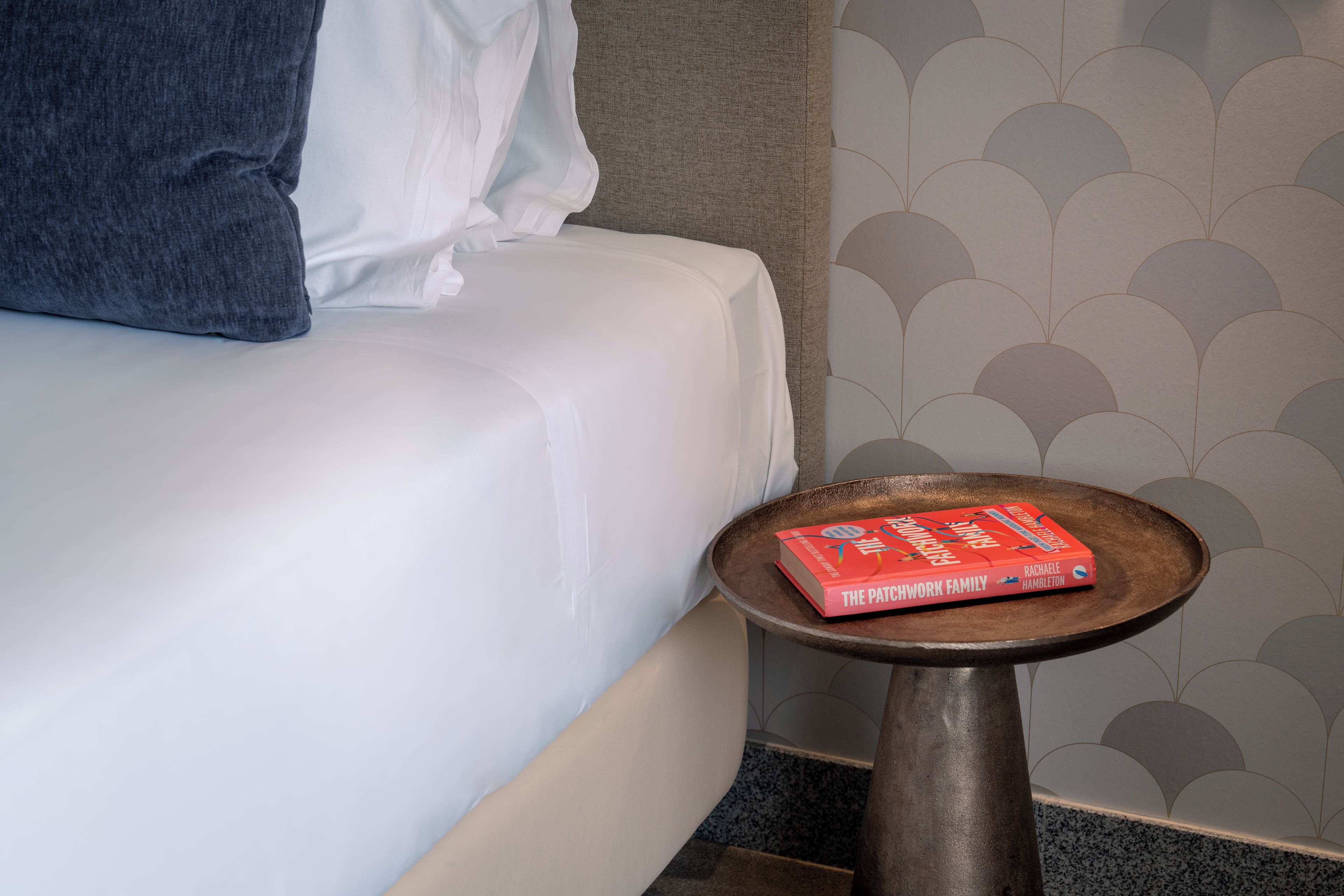A bed with a white sheet and a pillow, a small round table with a book on it, and a wall with a scalloped pattern.