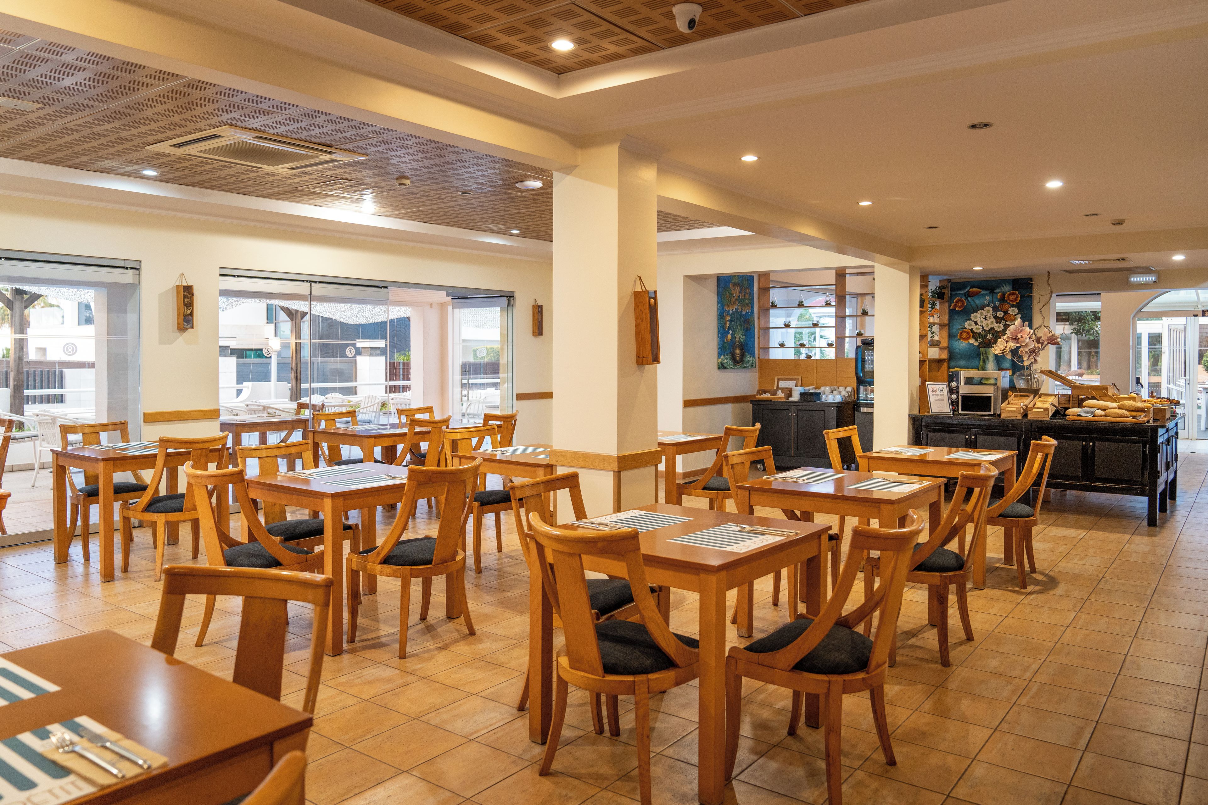 Un restaurant avec des tables et des chaises en bois, un espace cuisine et des œuvres d’art accrochées aux murs.