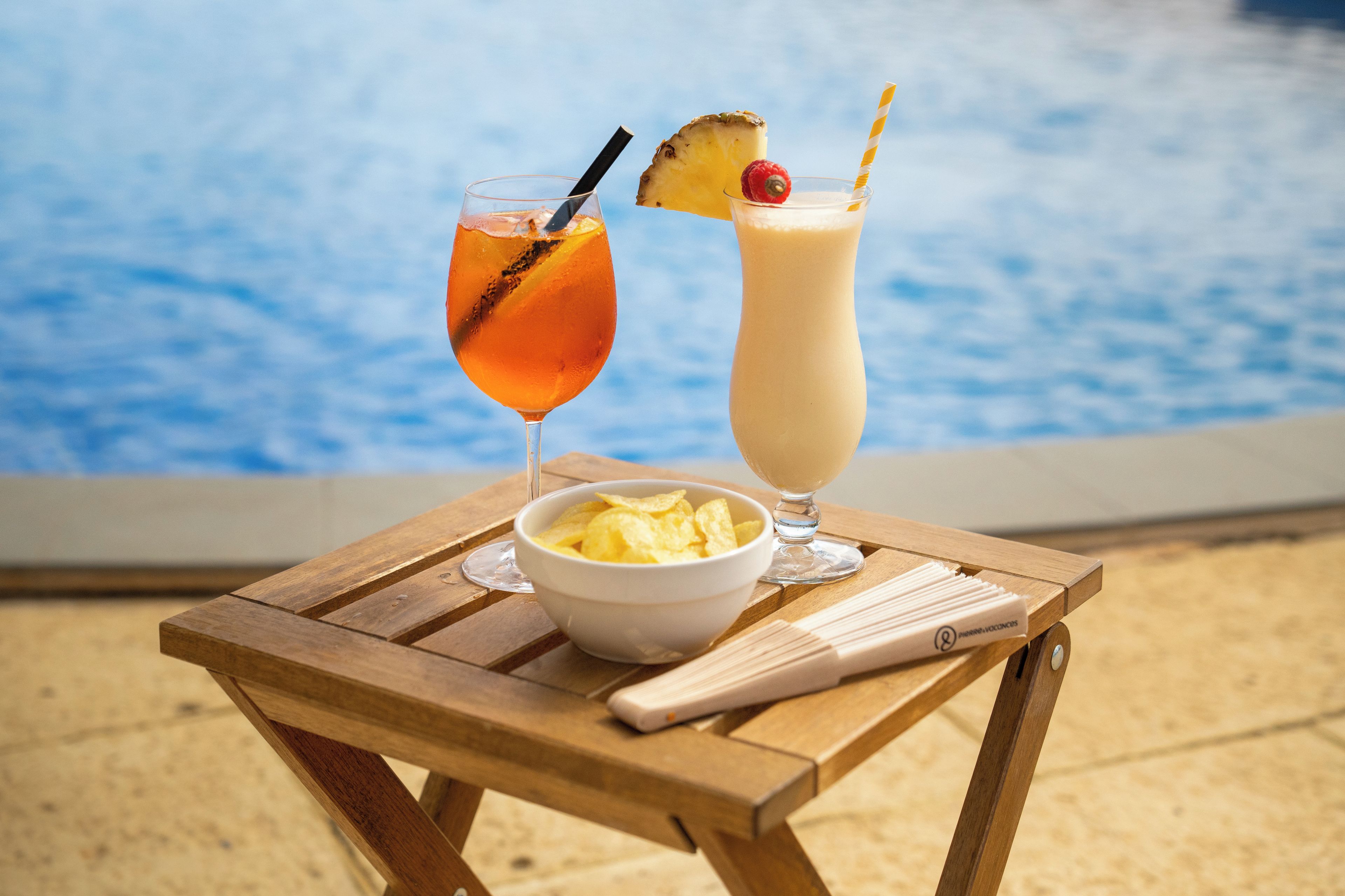 Ein Holztisch mit einer Schüssel Chips und zwei Getränken, eines mit Ananasstück und Trinkhalm, das andere mit Ananasstück und Himbeere, neben einem Pool.