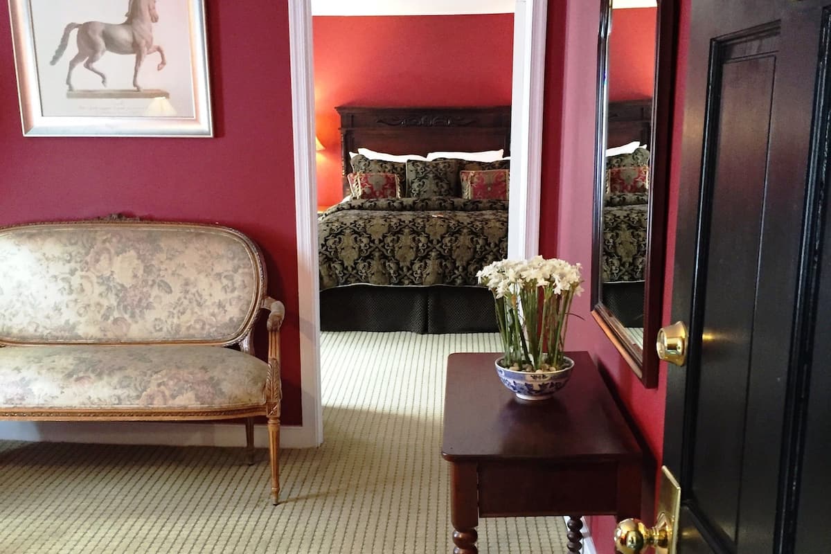 A room with a red wall, a framed horse picture, a sofa, a bedside table with a vase, and a bed with patterned bedding.