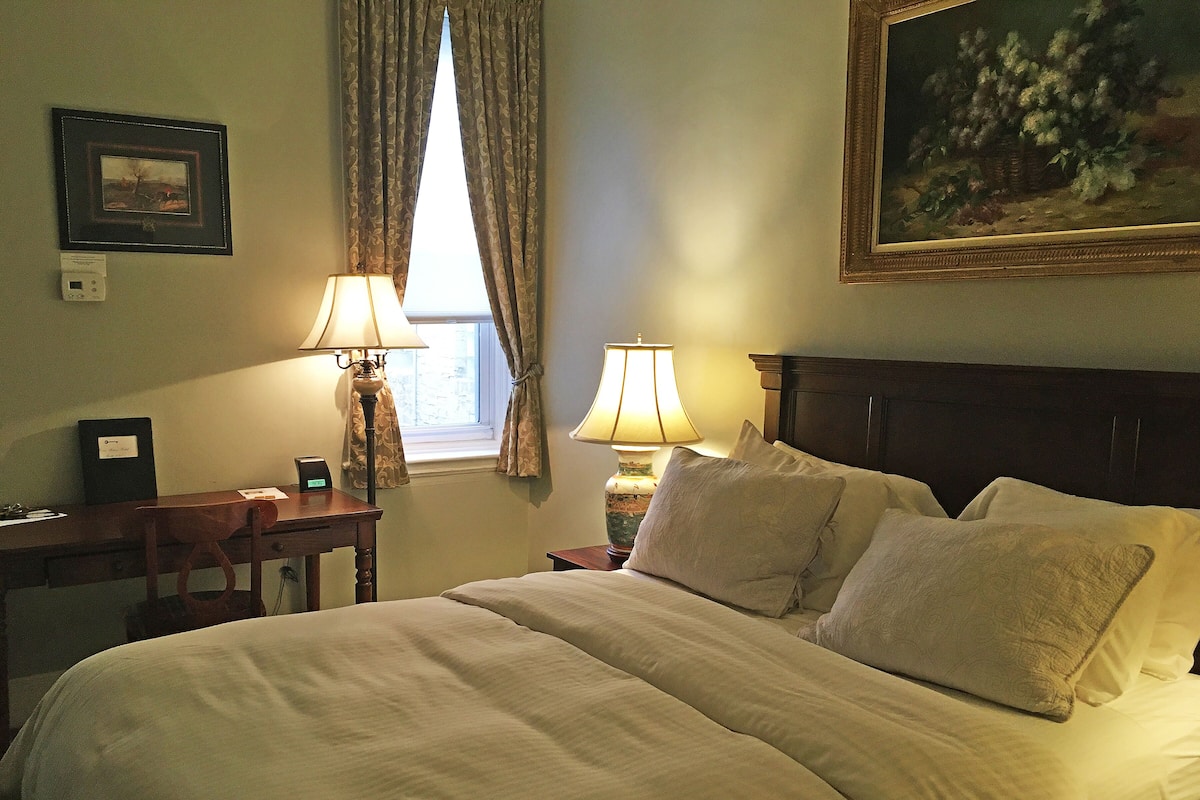 A neatly made bed with white linens, two bedside lamps, a wooden desk with a lamp, a framed picture on the wall, and a window with curtains.