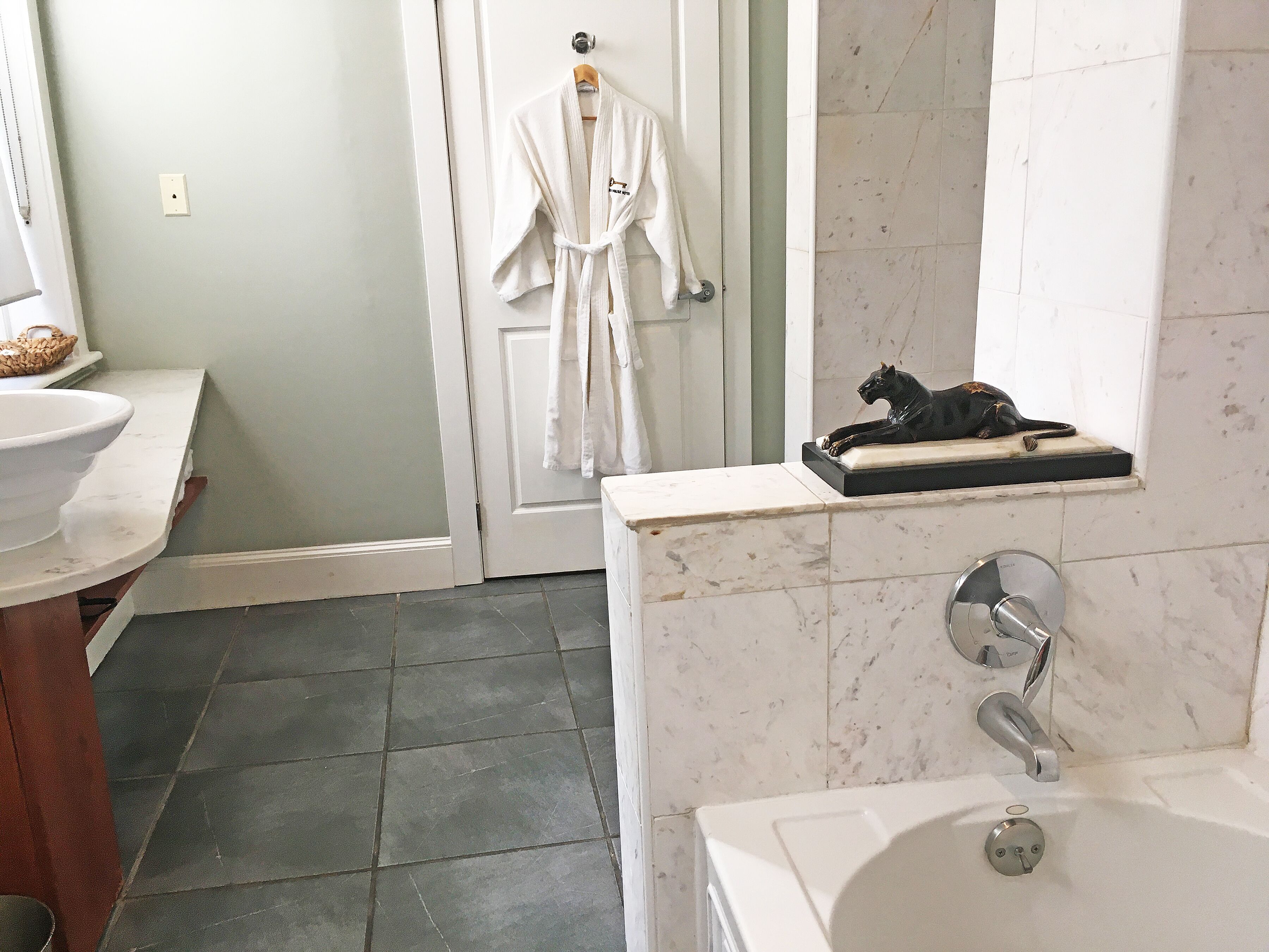 A bathroom with a bathtub, a sink, a mirror, a robe hanging on the door, and a statue on the countertop.