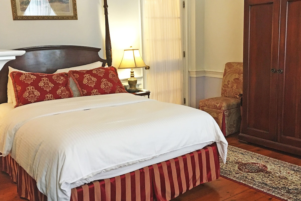 A neatly made bed with a white and red bedspread, a wooden headboard, and a red and white striped skirt. There is a wooden nightstand with a lamp, a chair, and a wooden wardrobe.