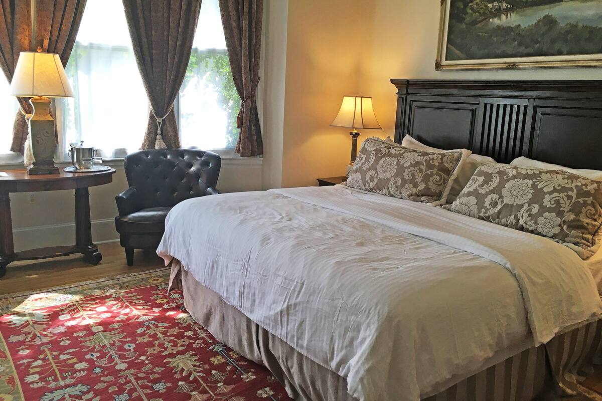 A neatly made bed with a floral-patterned pillow, a red patterned rug, and a painting above the bed.