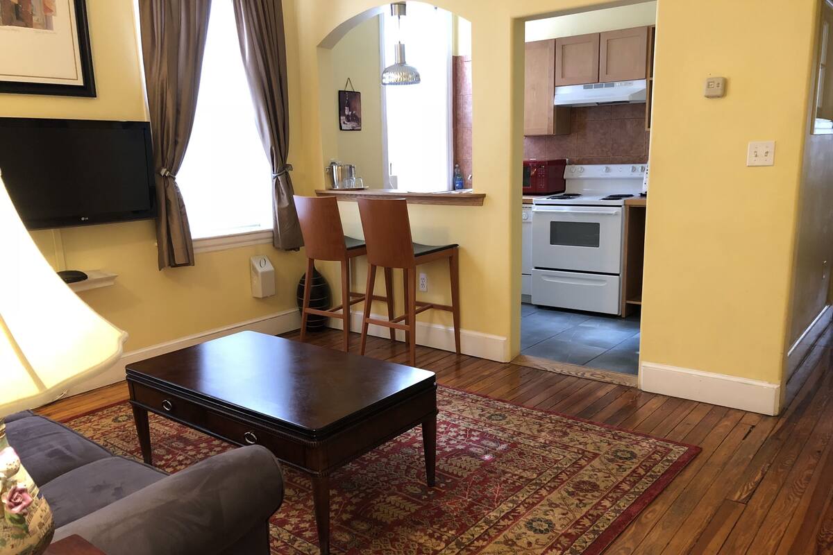 A living room with a dark wooden coffee table, a patterned rug, and a sofa. A kitchen is visible in the background with white appliances.
