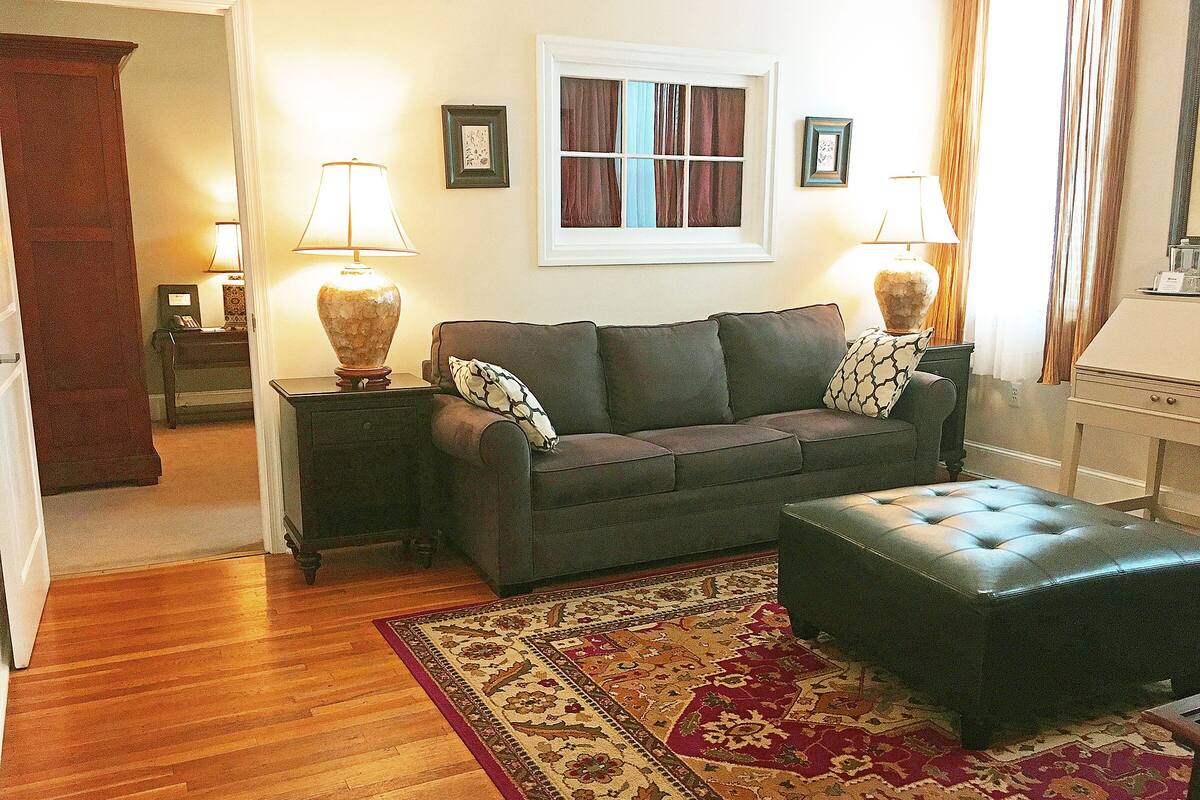 A living room with a sofa, ottoman, patterned rug, two lamps, and a window with curtains.