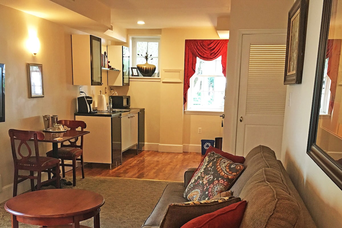 A cozy living room with a sofa, a coffee table, and a dining area with chairs and a small kitchen in the background.