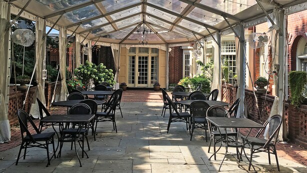 A covered patio with tables and chairs, a glass ceiling, and a brick building in the background.