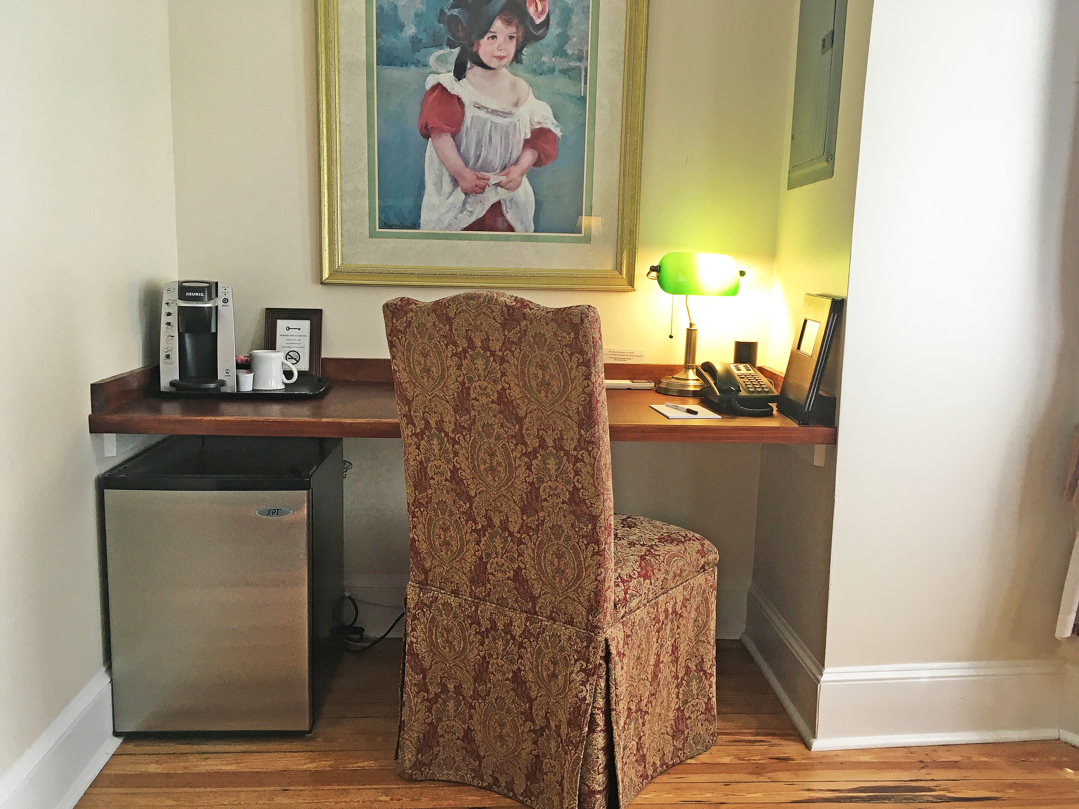 A hotel room with a desk, a coffee machine, a telephone, a small refrigerator, and a patterned chair.