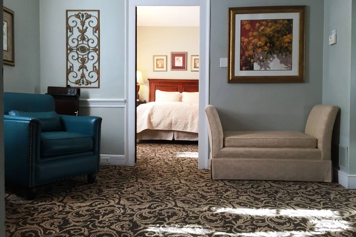 A hotel room with a patterned carpet, a blue armchair, a beige armchair, a bed with a white duvet, and a framed painting on the wall.