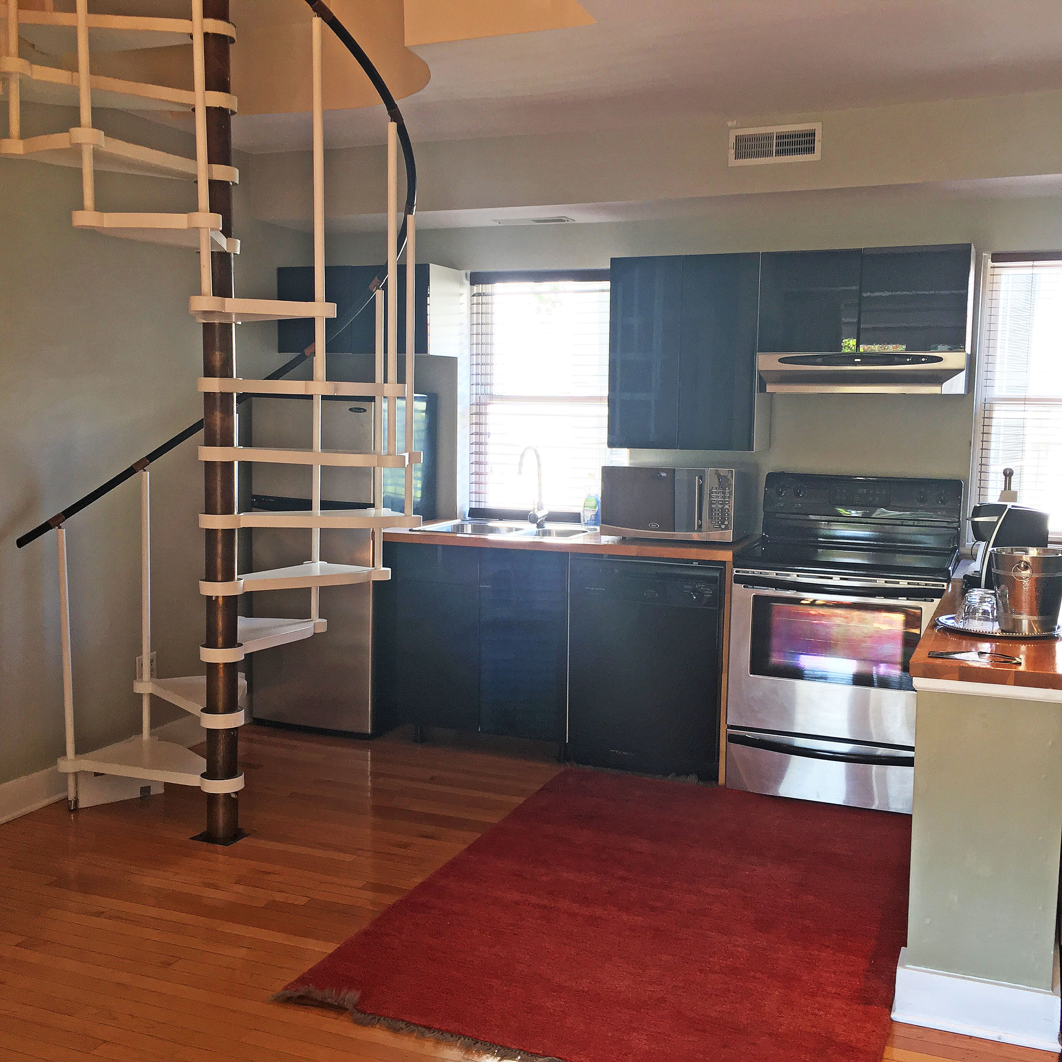 A kitchen with a built-in oven, microwave, and dishwasher. A red rug is on the floor, and a spiral staircase is visible.