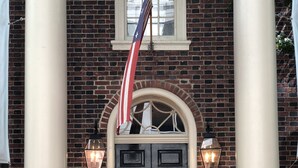A brick building with a black double door, two white columns, and an American flag.