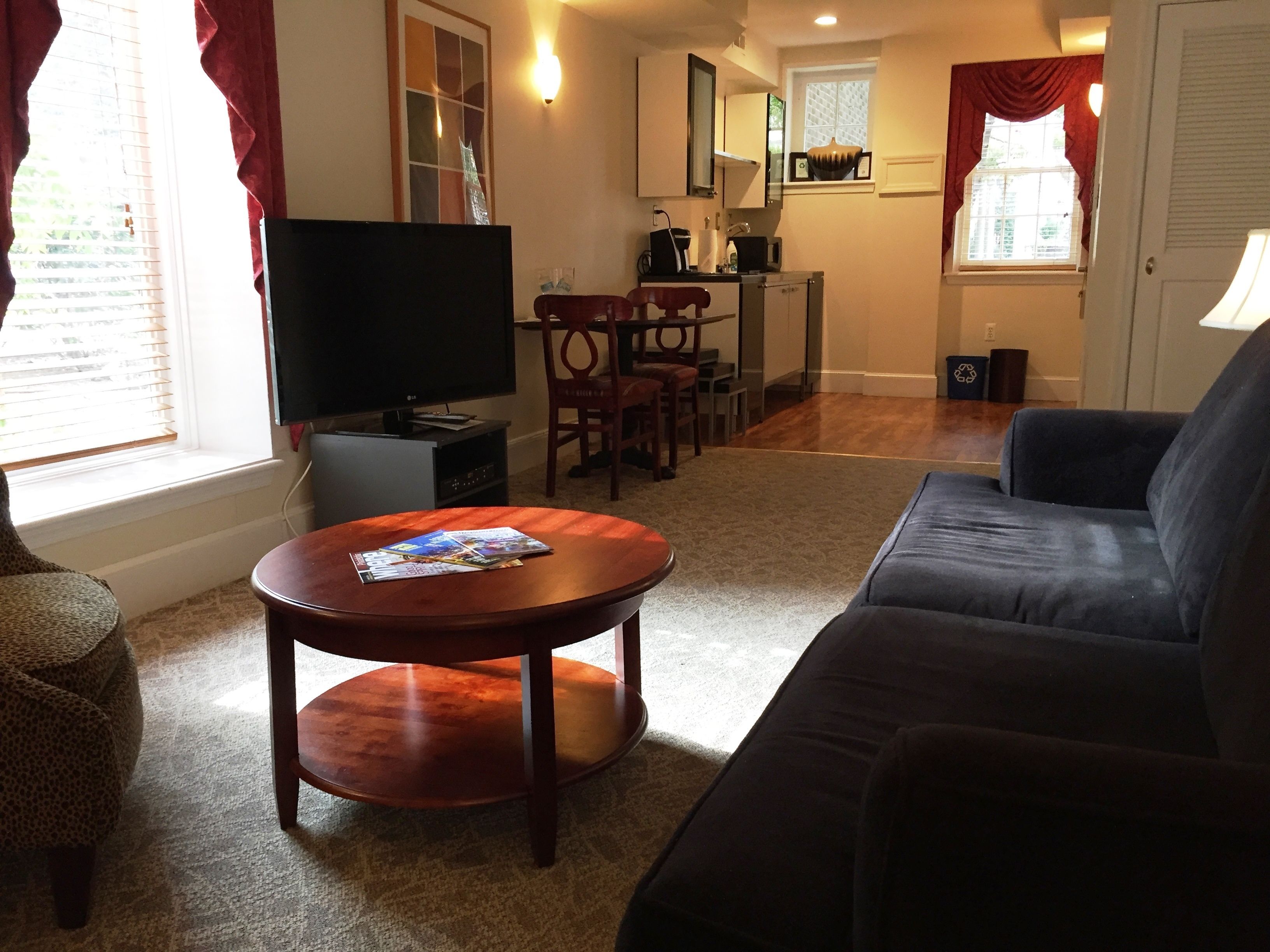A living room with a dark-colored sofa, a wooden coffee table with magazines, a television, and a dining area with a table and chairs.