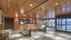 Un vestíbulo moderno con paneles de techo de madera, un mostrador de recepción, un área de estar con sofás y una mesa de centro, y grandes ventanales con vistas a la calle.