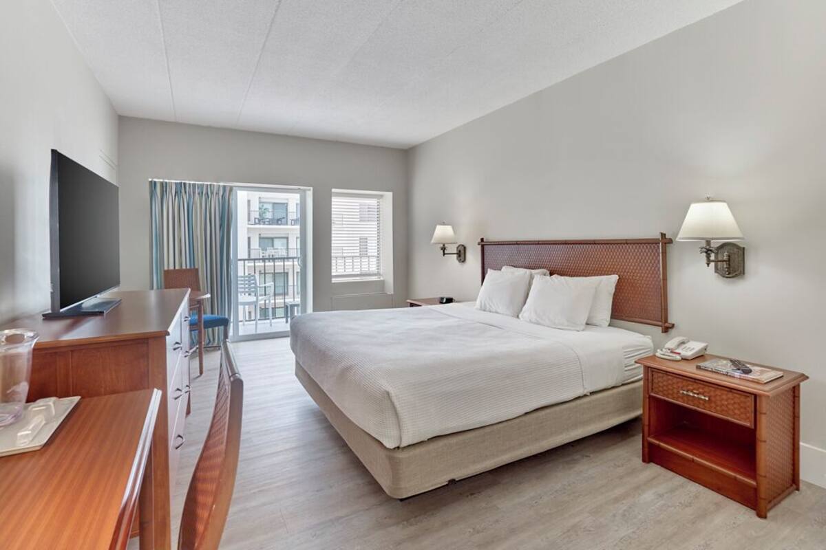 Habitación de hotel con una cama grande, un escritorio con silla, un televisor, una mesita de noche con lámpara y una ventana con persianas.