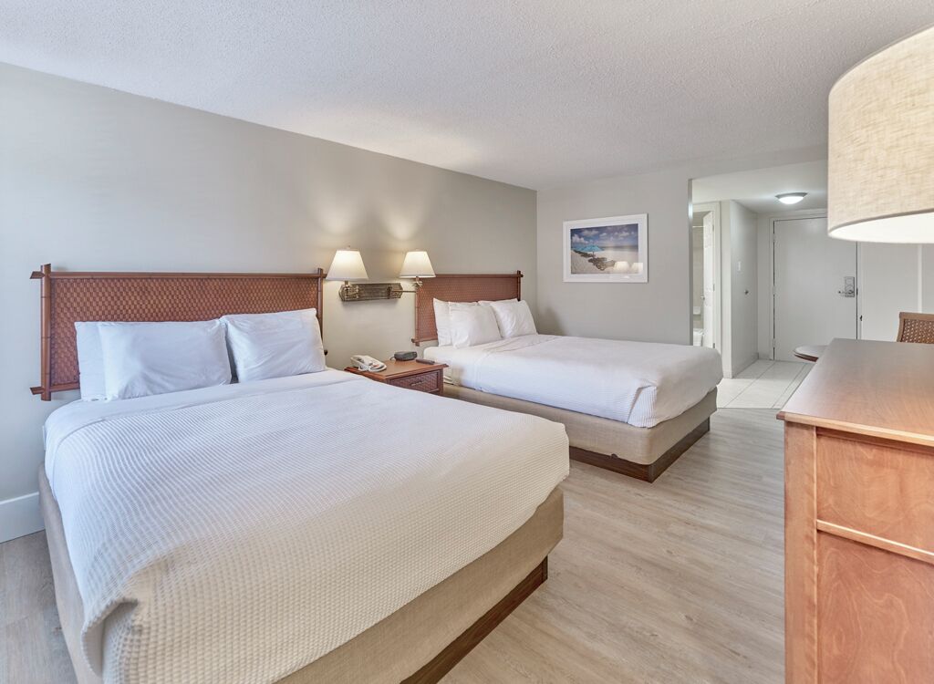 A hotel room with two beds, a wooden headboard, bedside lamps, a nightstand, and a desk with a lamp.