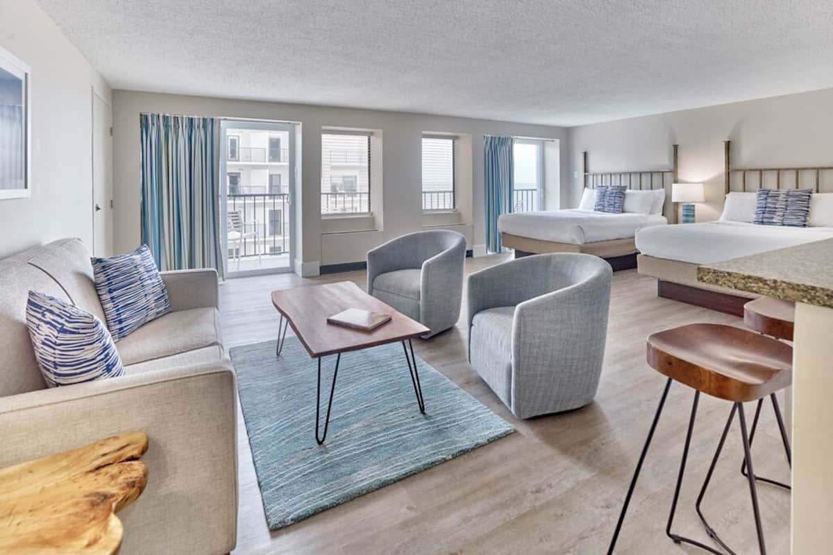 Habitación de hotel moderna con sofá, dos sillones, una mesita, cama, zona de bar y ventanas grandes con cortinas.