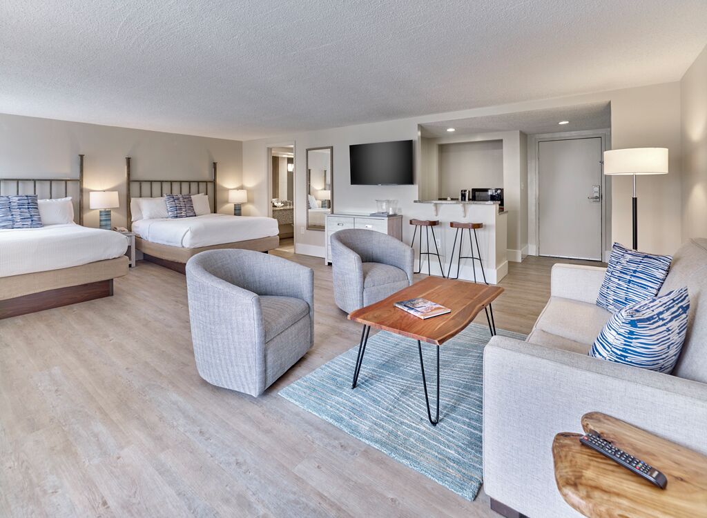 A hotel room with two beds, a sofa, a coffee table, a TV, a kitchen area, and a bathroom.