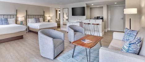 Habitación de hotel con dos camas, un sofá, una mesa de centro, un televisor, una zona de cocina y un baño.
