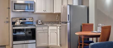 Cocina compacta con armarios blancos, microondas de acero inoxidable, horno y refrigerador. Hay una mesa de comedor con sillas y un sofá azul en la sala.