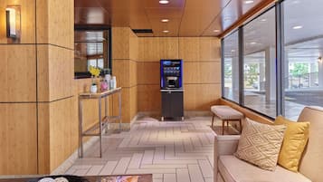Un hall d’hôtel avec un décor intérieur en bois, un espace salon avec un canapé et des coussins, une petite table agrémentée d’objets décoratifs et un kiosque en libre-service.