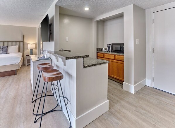 A compact hotel room with a kitchenette, a bed with a headboard, a desk with a lamp, and a TV mounted on the wall.