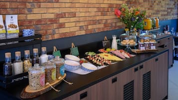 Un mostrador de buffet con diversas opciones de comida, entre ellas frutas, quesos y panes, sobre un fondo de pared de ladrillo.