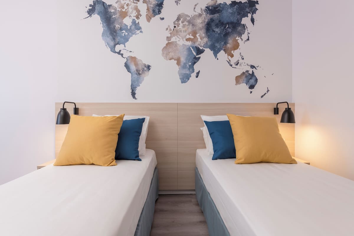 Une chambre d’hôtel avec deux lits, chacun agrémenté d’un oreiller jaune et bleu, et une carte du monde accrochée au mur.