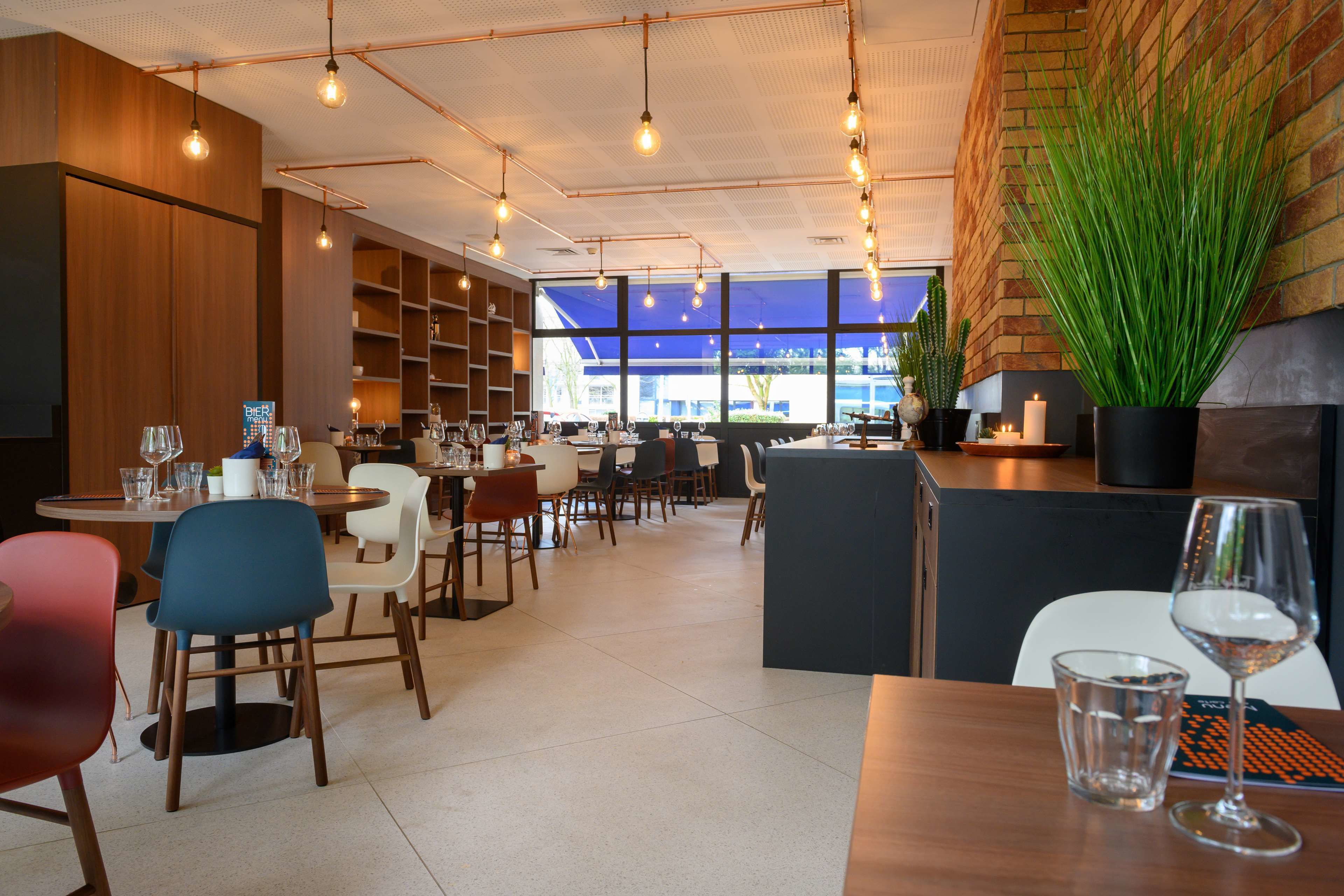 A modern restaurant with wooden walls, a brick accent wall, and a large potted plant. Tables are set with wine glasses and cutlery.