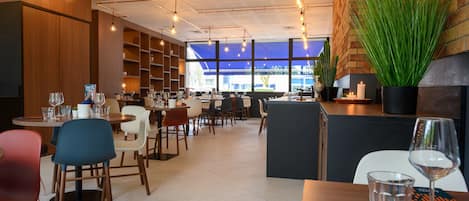 Un restaurante moderno con paredes de madera, un detalle de pared de ladrillo y una planta grande en maceta. Las mesas están dispuestas con copas de vino y cubiertos.
